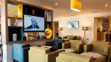 Un área de descanso moderna con asientos cómodos, un televisor mostrando noticias y una zona de bar con estantes y objetos decorativos.