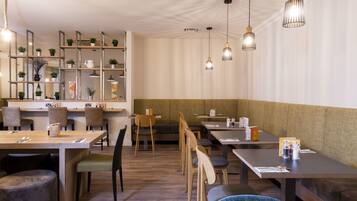 A modern restaurant with wooden tables and chairs, pendant lighting, and shelves with plants and kitchenware.