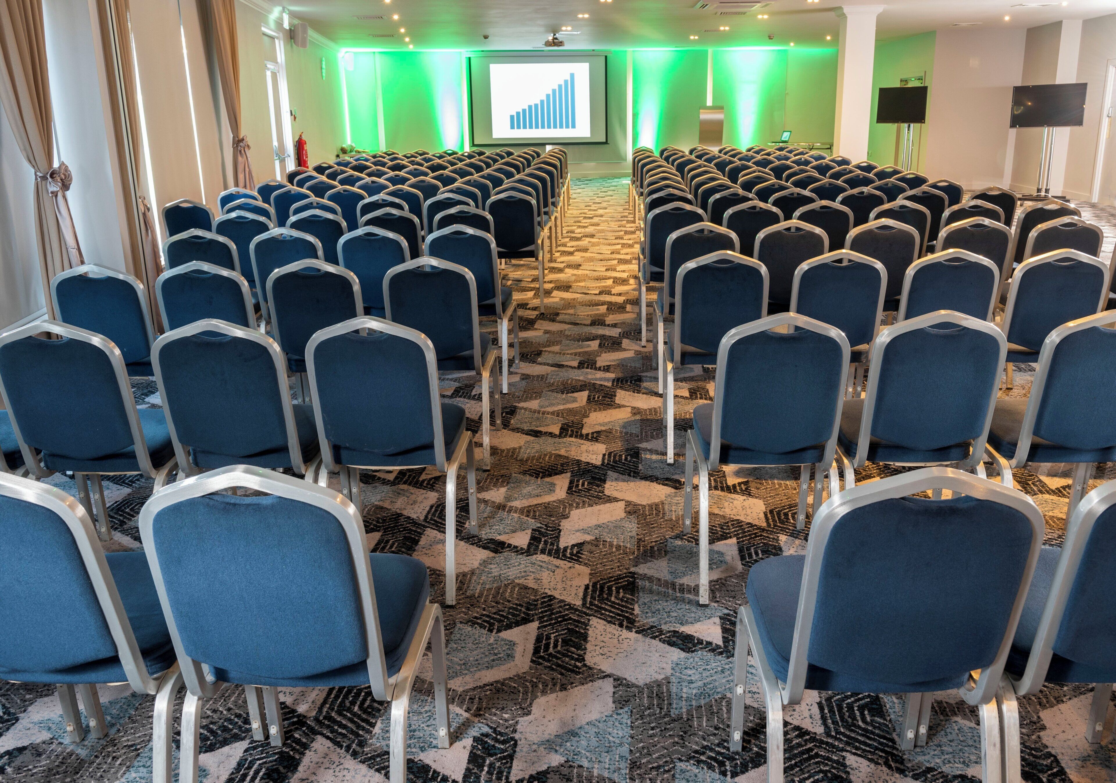 Un salón de conferencias con filas de sillas, piso alfombrado y una pantalla para presentaciones.