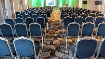 Un salón de conferencias con filas de sillas, piso alfombrado y una pantalla para presentaciones.