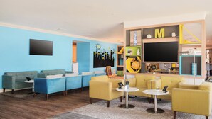 Un moderno vestíbulo de hotel con una decoración azul y amarilla, que incluye una zona de estar con sofás y sillones, un televisor de pantalla plana y un área de juegos infantiles con un globo terráqueo y juguetes educativos.