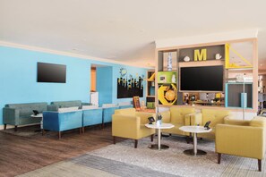 Un moderno vestíbulo de hotel con una decoración azul y amarilla, que incluye una zona de estar con sofás y sillones, un televisor de pantalla plana y un área de juegos infantiles con un globo terráqueo y juguetes educativos.