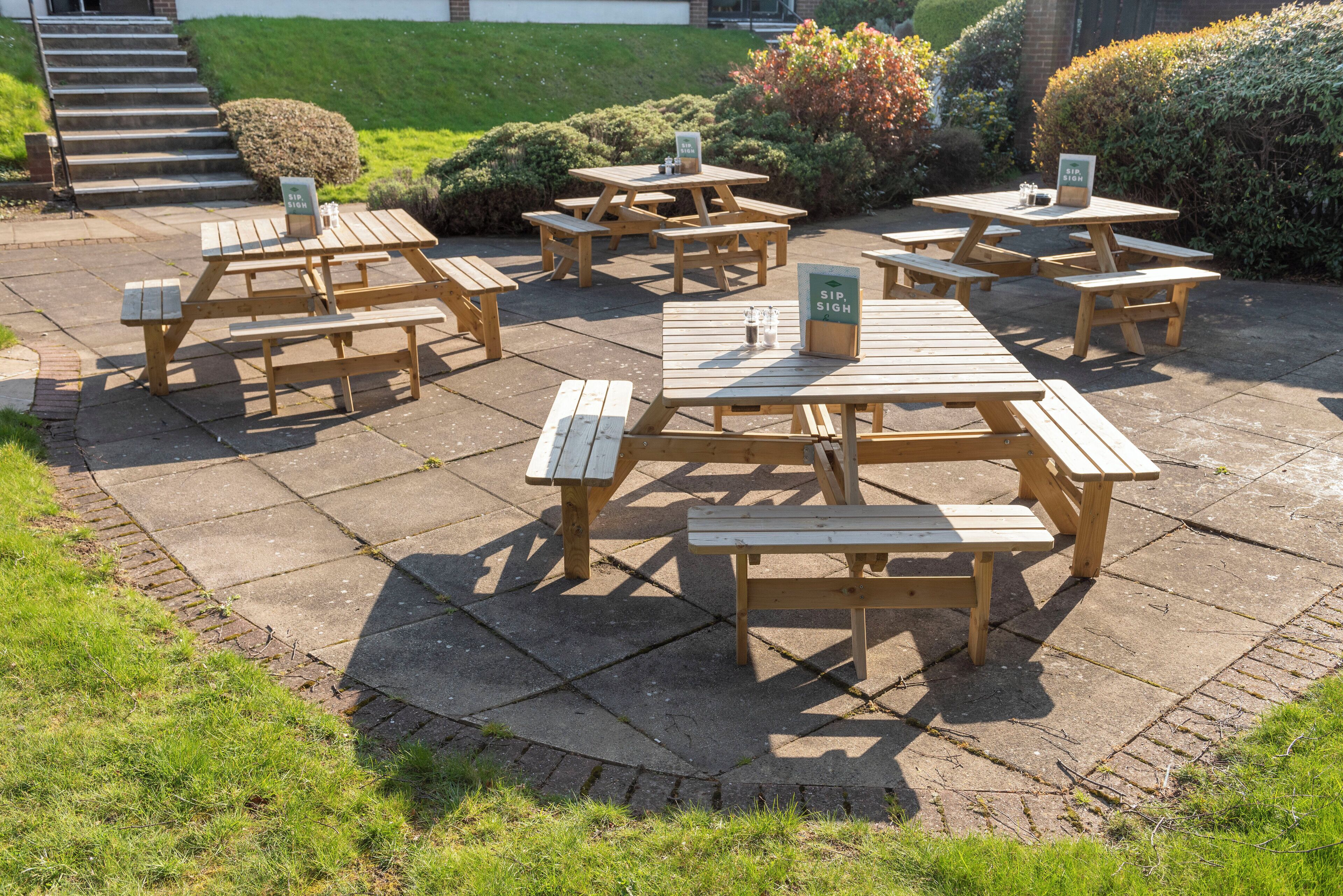 An outdoor seating area with multiple wooden picnic tables and benches, arranged on a paved surface.