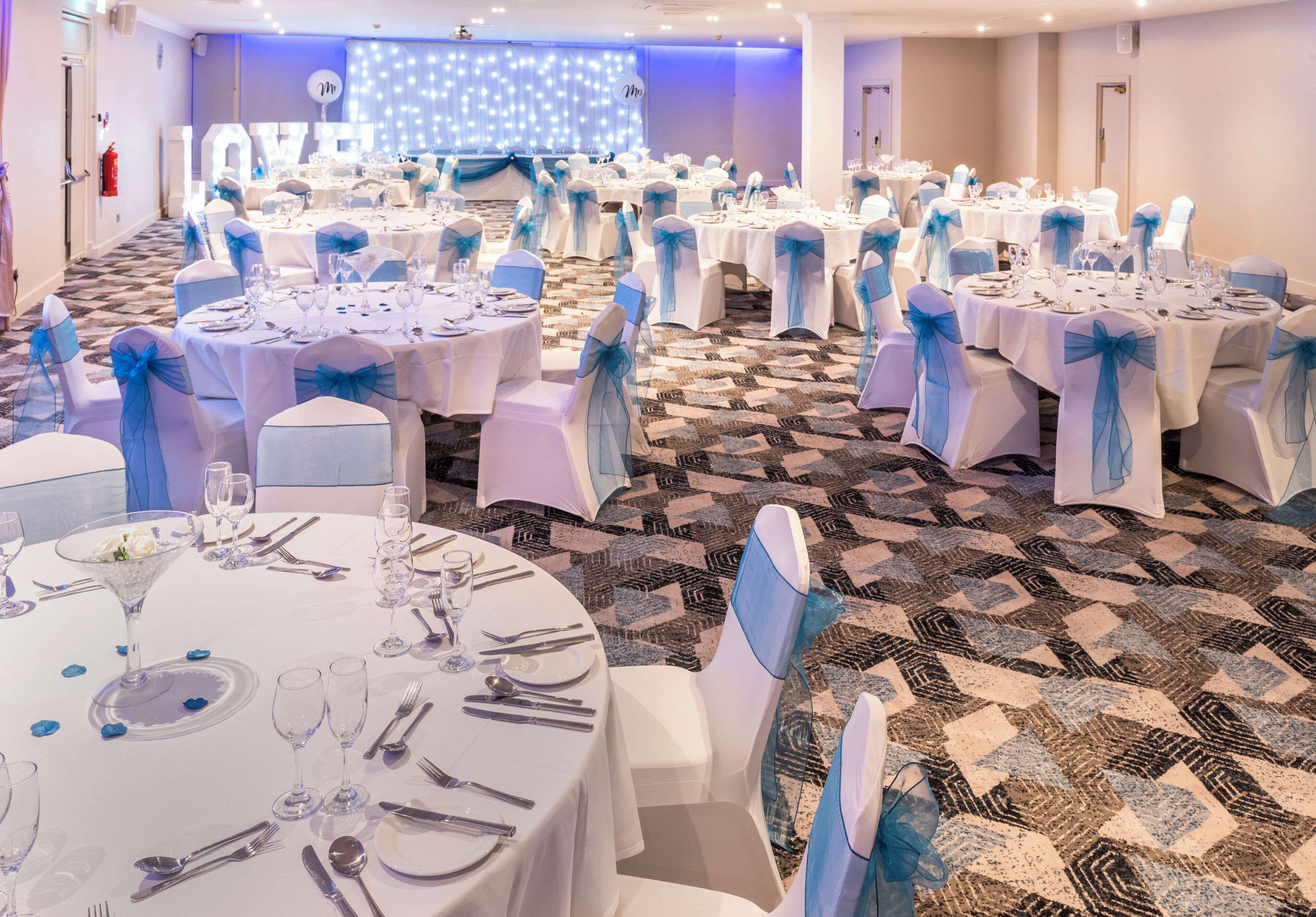 A banquet hall with round tables set for dining, chairs with blue sashes, and a stage with a light backdrop.
