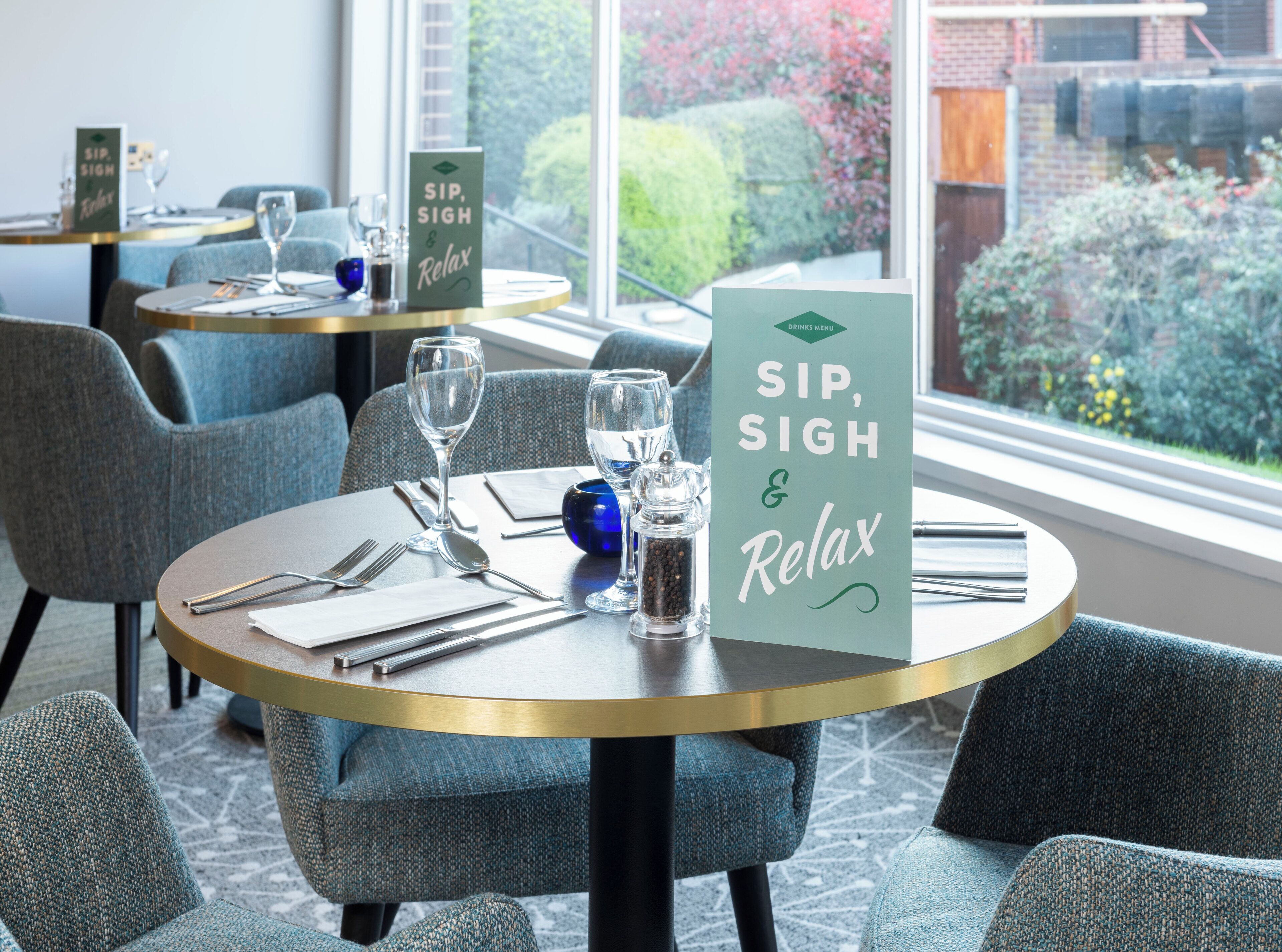 A dining area with round tables, chairs, and a menu stand displaying 'SIP, SIGH & Relax'.