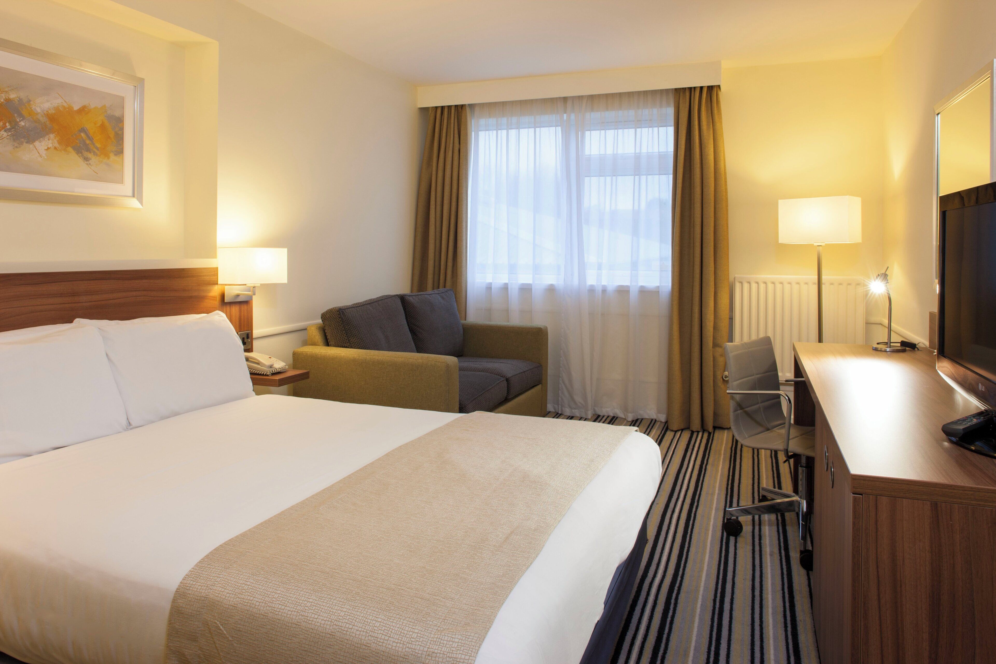 Una habitación de hotel con una cama grande, un escritorio con una silla, un televisor y una ventana con cortinas.