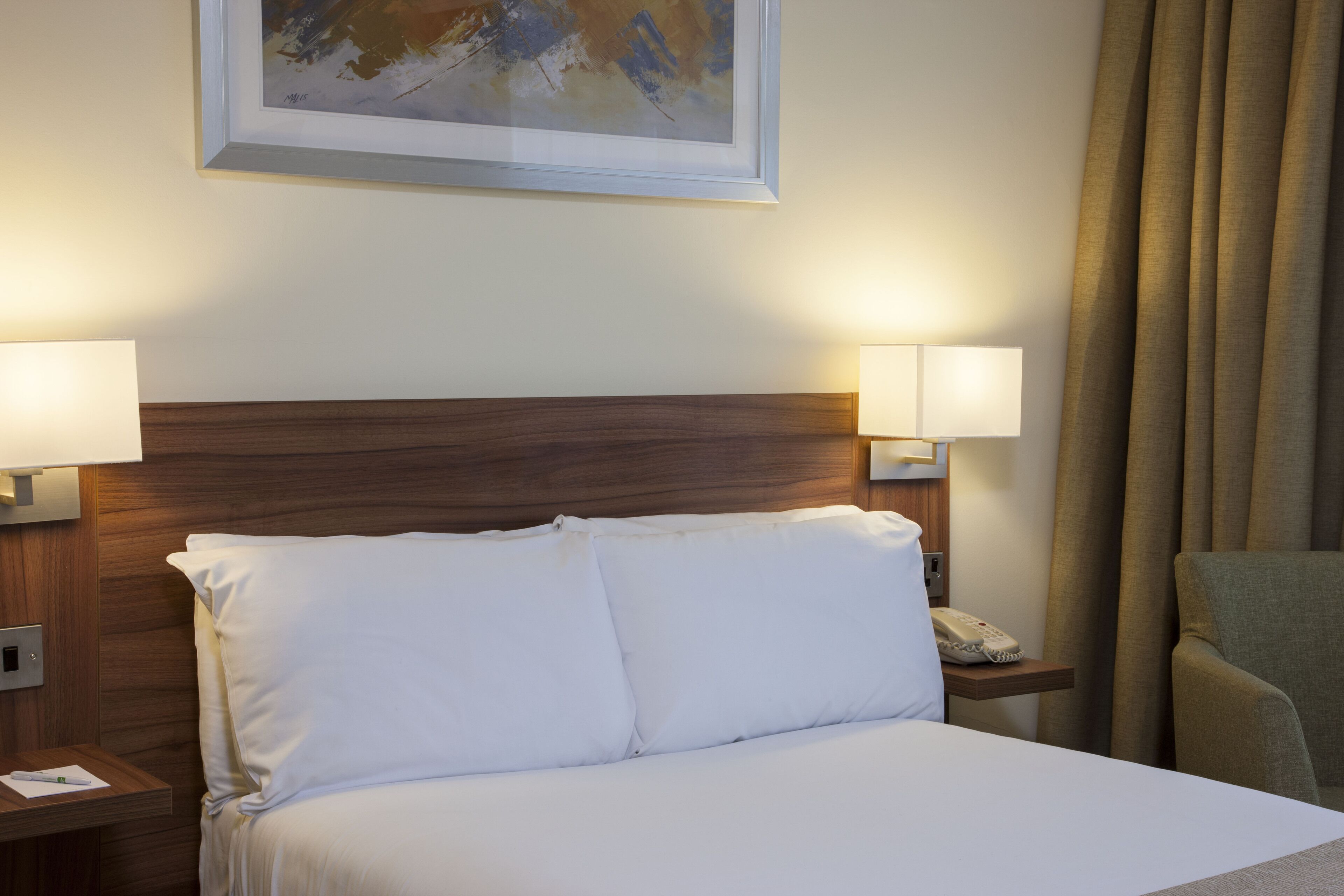 A hotel room with a wooden headboard, white bedding, two bedside lamps, a telephone, and a framed artwork on the wall.