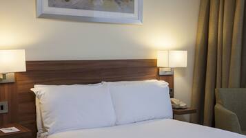 A hotel room with a wooden headboard, white bedding, two bedside lamps, a telephone, and a framed artwork on the wall.