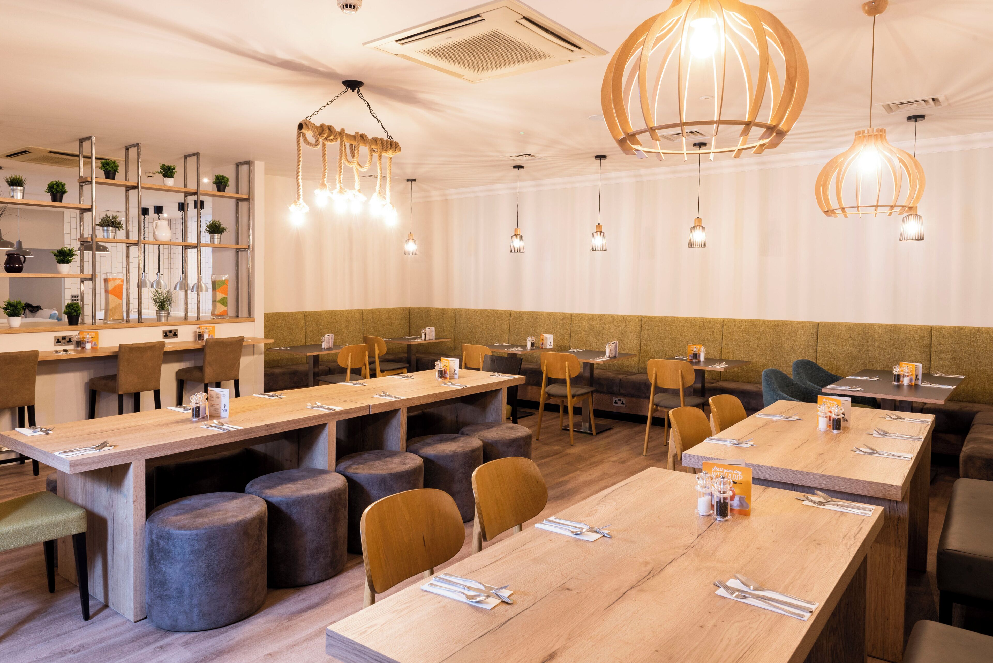 A restaurant with long wooden tables, cushioned seating, pendant lighting, and a neutral color palette.
