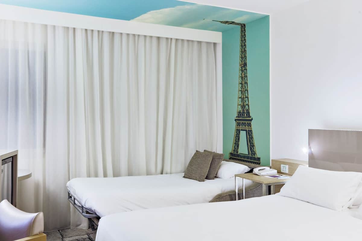 Ein Hotelzimmer mit zwei Betten, einem großen Fenster mit weißen Vorhängen, einem Wandbild mit dem Eiffelturm und einem Nachttisch mit Lampe und Telefon.