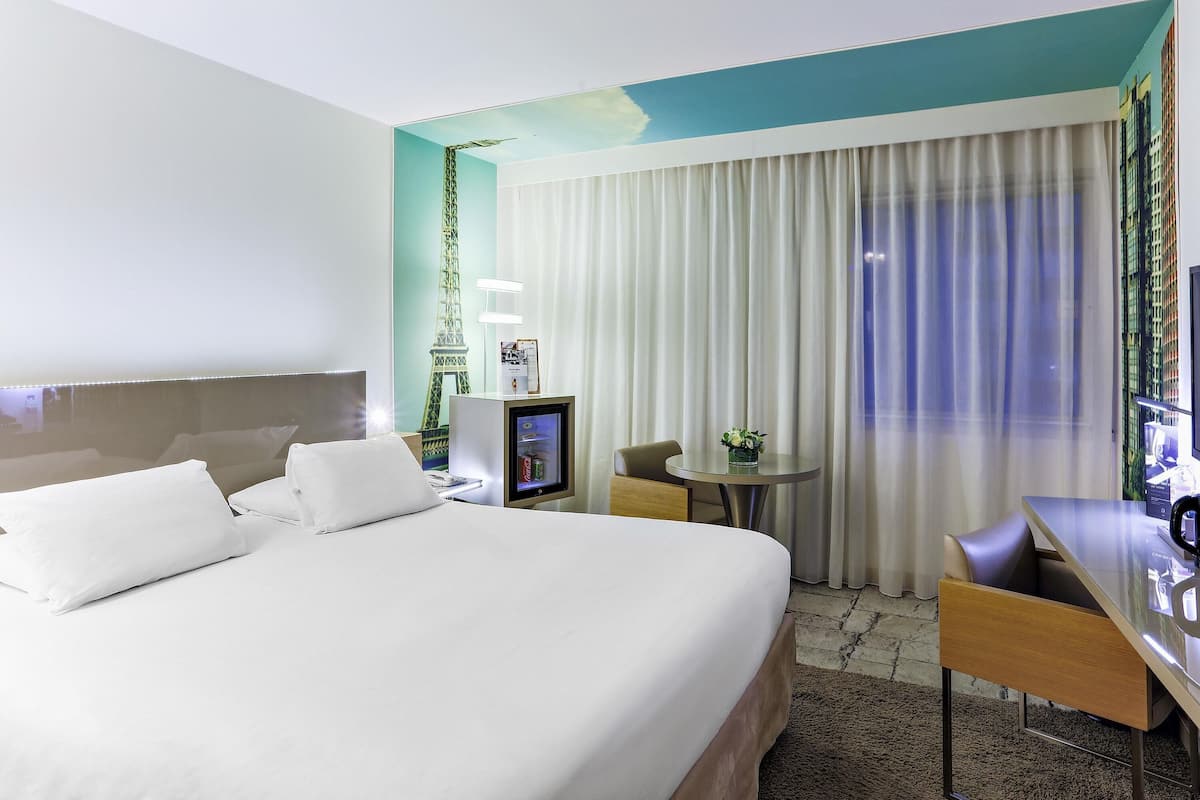 Ein Hotelzimmer mit Bett, Schreibtisch, Stuhl, einem kleinen Tisch mit Vase, Fernseher und Vorhang.