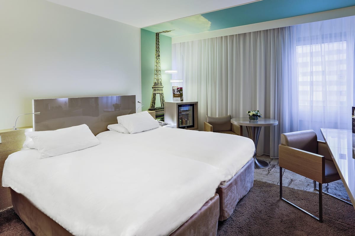 Ein Hotelzimmer mit Bett, Schreibtisch, Stuhl, Minibar und einem großen Fenster mit Vorhängen.