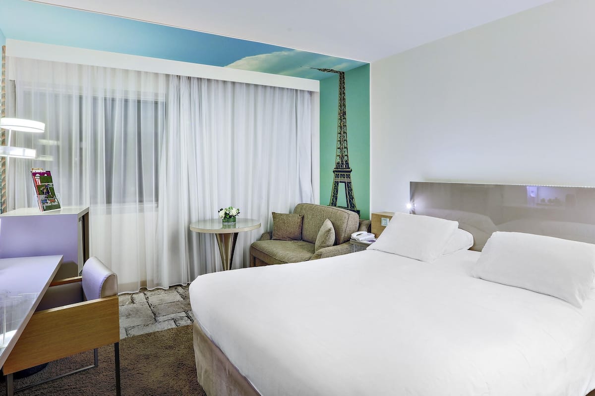 Ein Hotelzimmer mit Bett, Schreibtisch, Stuhl, einem kleinen Tisch mit Blumenvase und einer Wand mit Wandgemälde vom Eiffelturm.