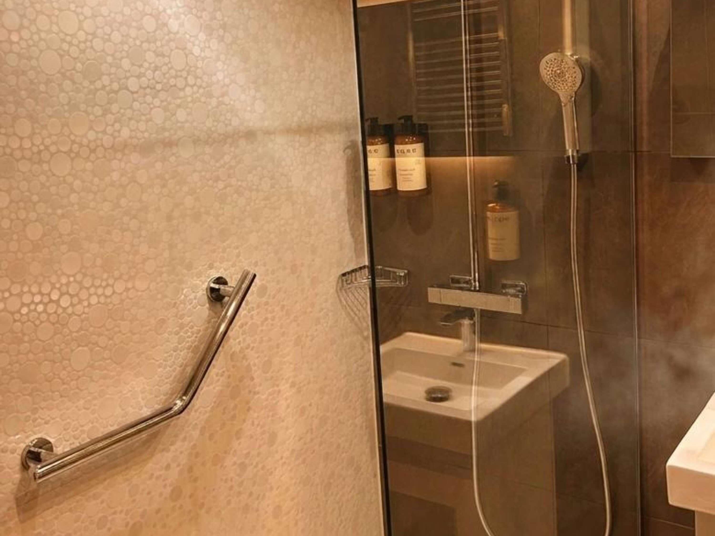 A bathroom with a glass shower enclosure, a handrail, and a sink.