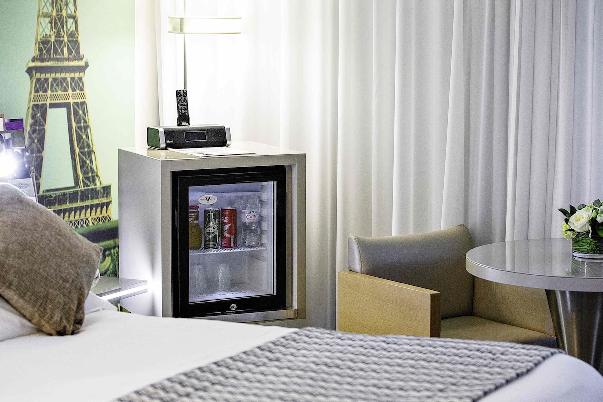 Ein Hotelzimmer mit Bett, Minibar, einem kleinen Tisch mit einer Vase und einer Wand mit Wandgemälde.