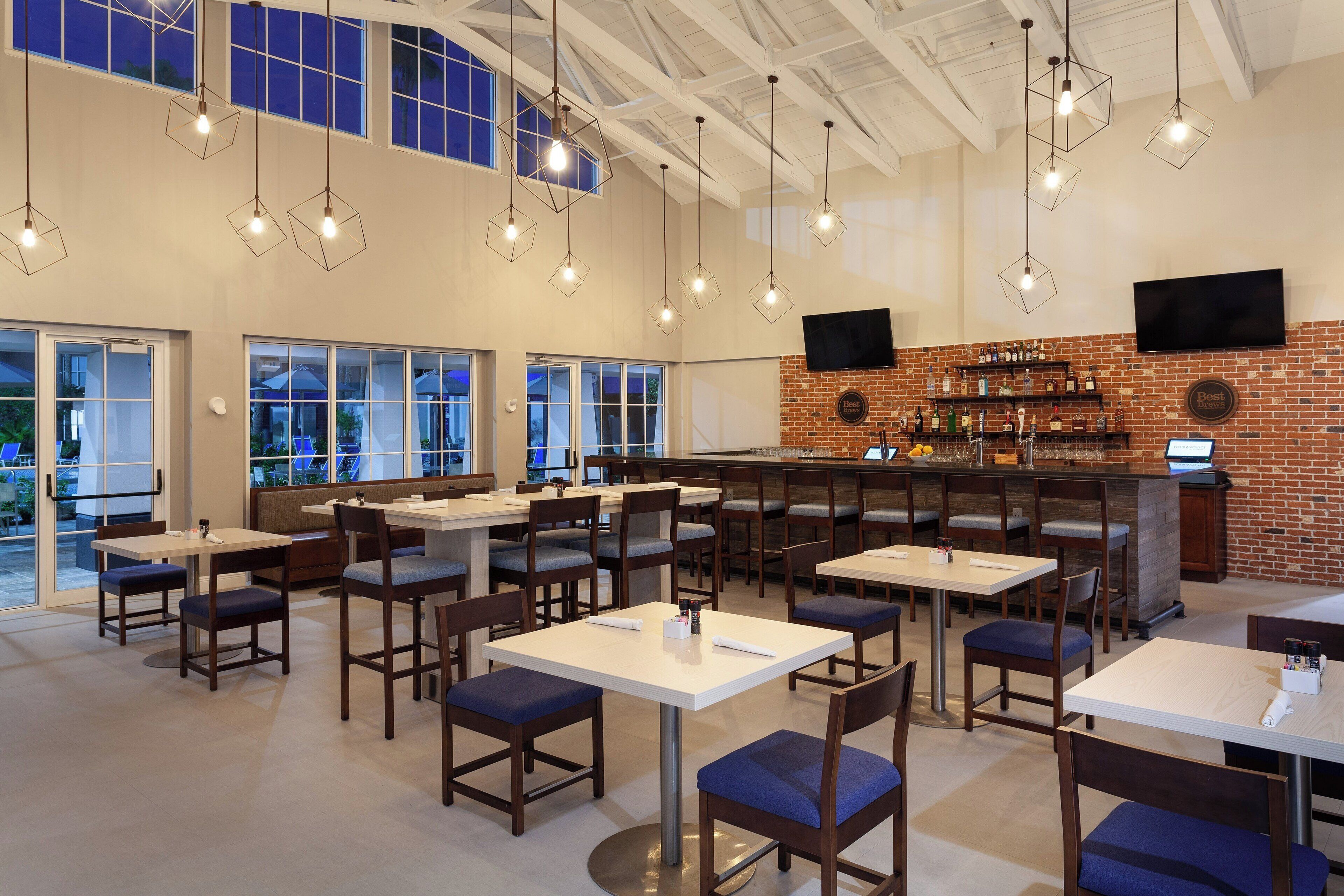 A restaurant with multiple tables and chairs, a brick wall with a bar area, and a large window overlooking the outside.