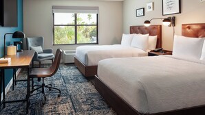A hotel room with two beds, a desk, a chair, a large window with a view of palm trees, and framed pictures on the wall.