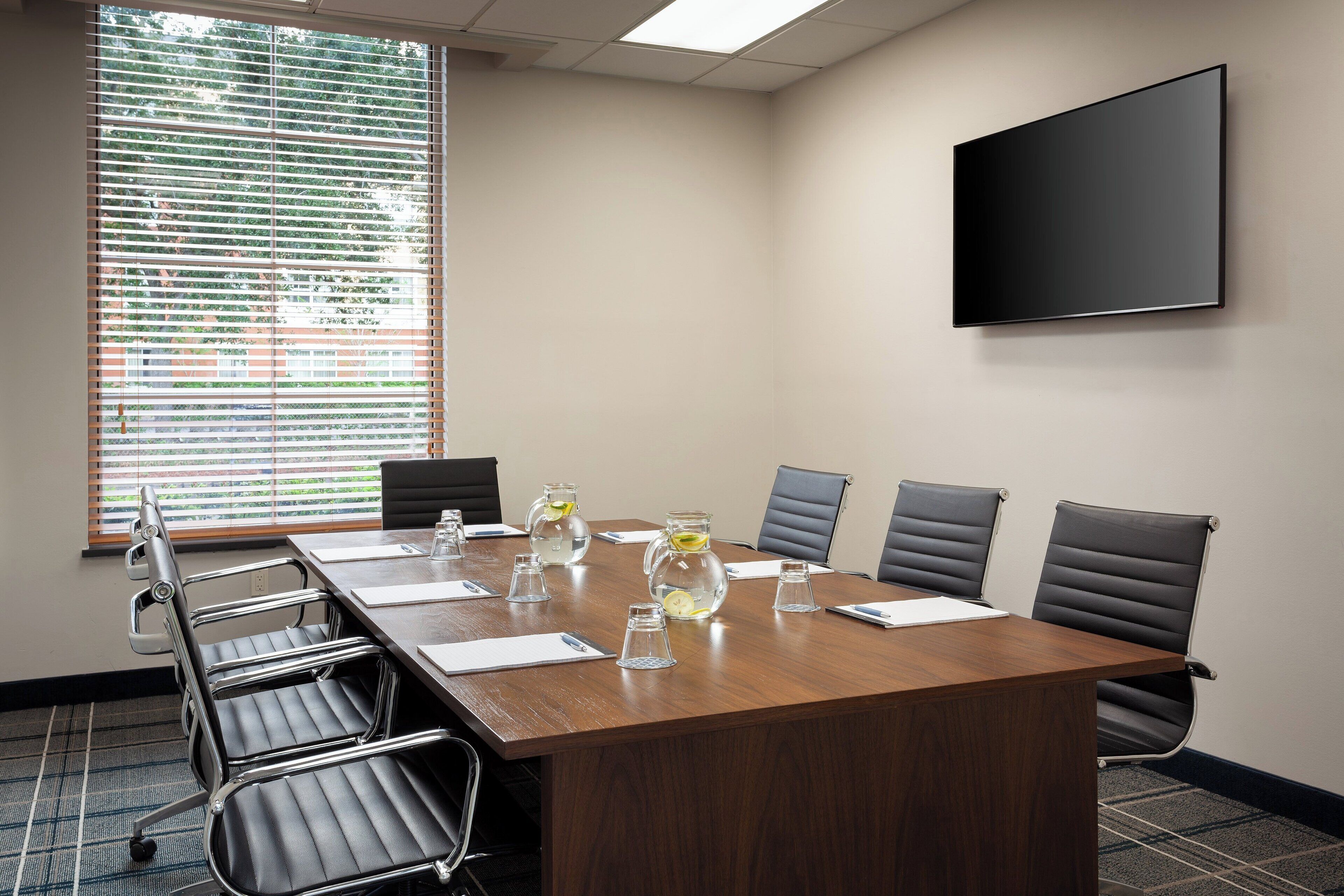 A conference room with a long table, six chairs, a flat-screen TV, and a window with blinds.