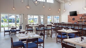 A spacious dining area with multiple tables and chairs, large windows offering views of the outdoors, and a bar area with a brick wall and various bottles.