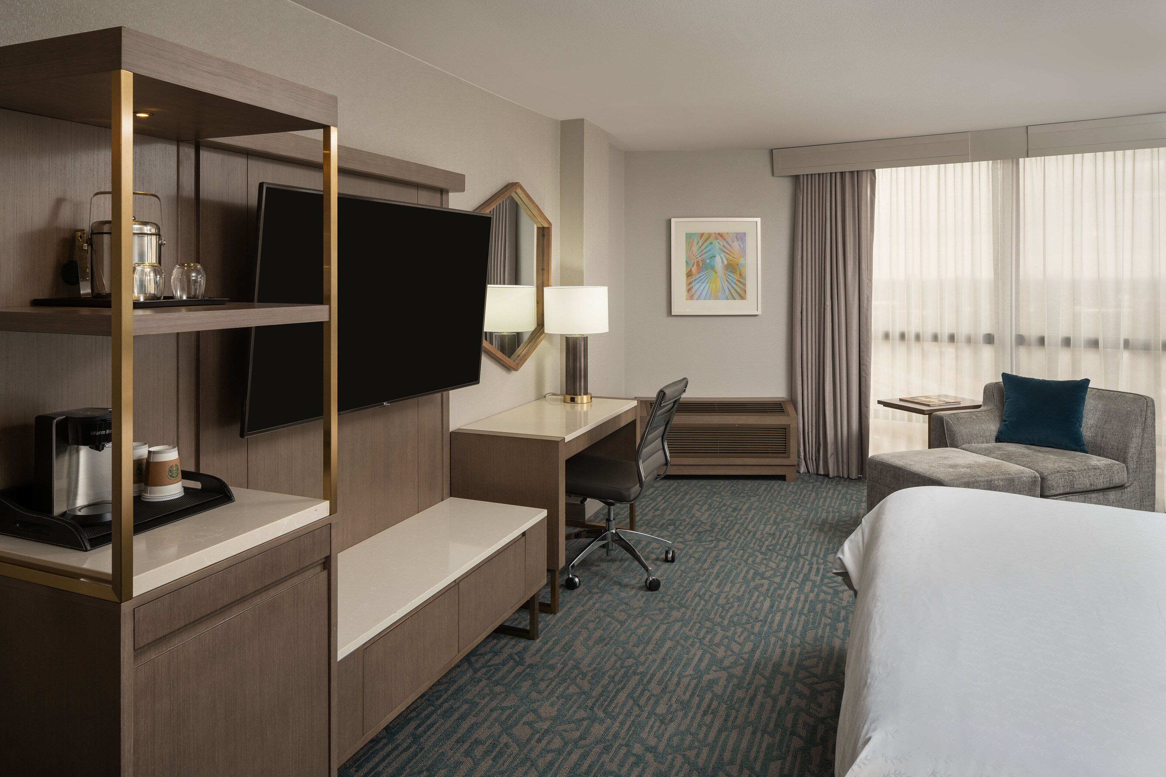 A hotel room with a bed, desk, chair, TV, coffee maker, and a window with curtains.