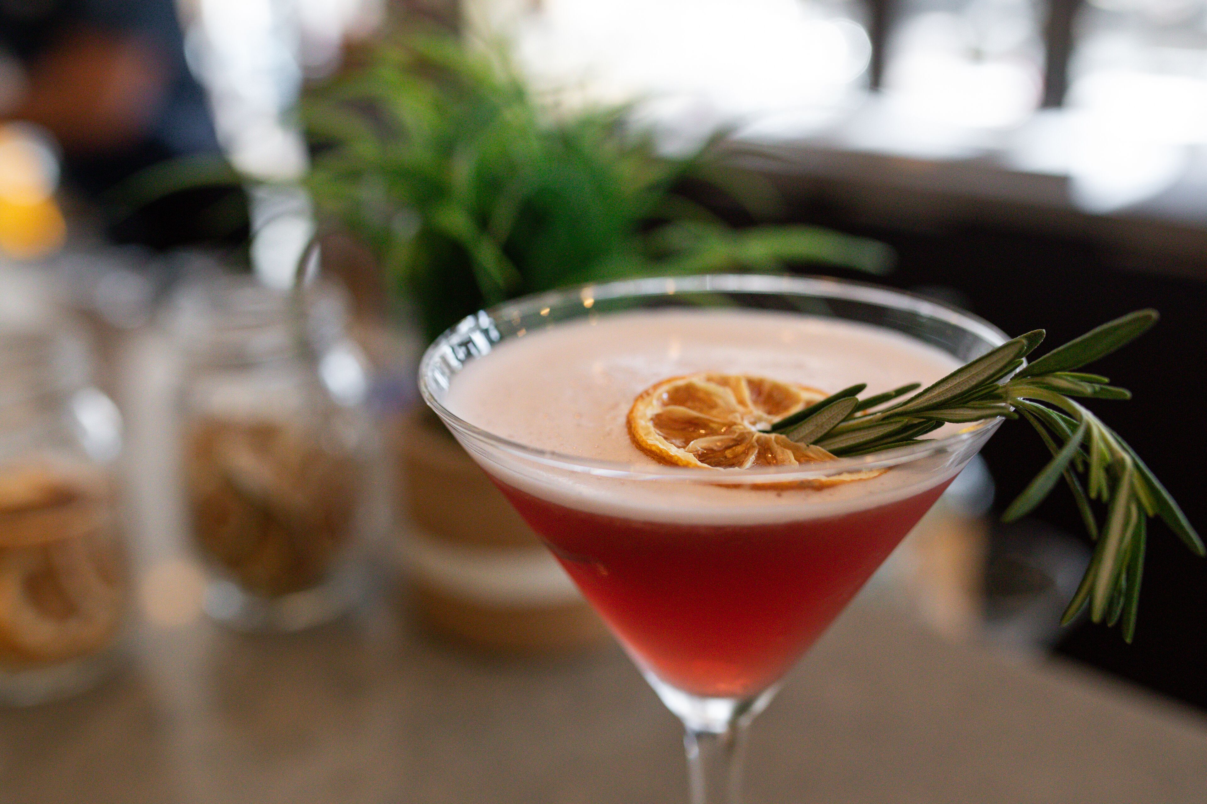 Un bicchiere da Martini con un liquido rosso, guarnito con una fetta d'arancia e un rametto di rosmarino.