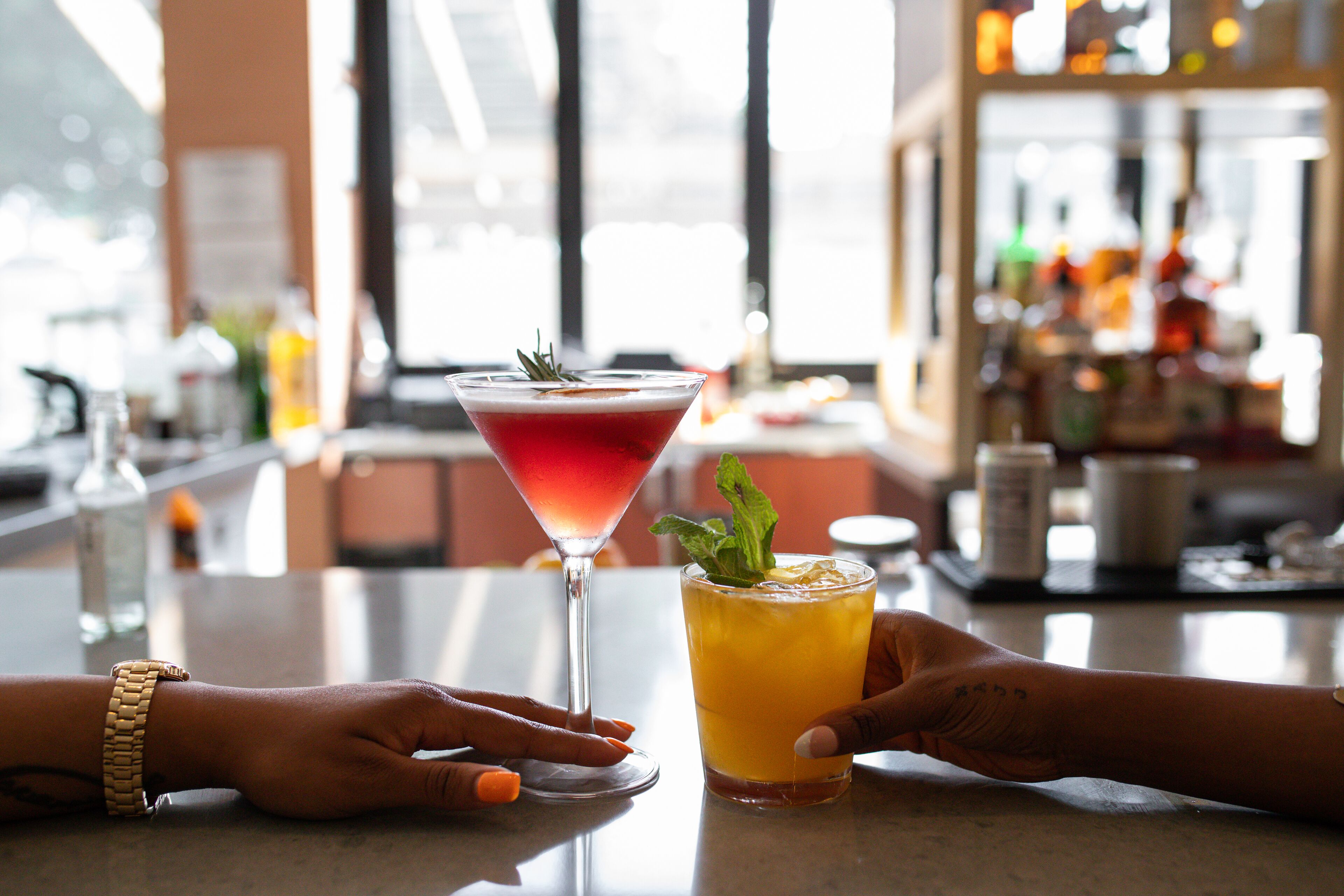 A bar setting with two cocktails on a table, one in a martini glass and the other in a highball glass.