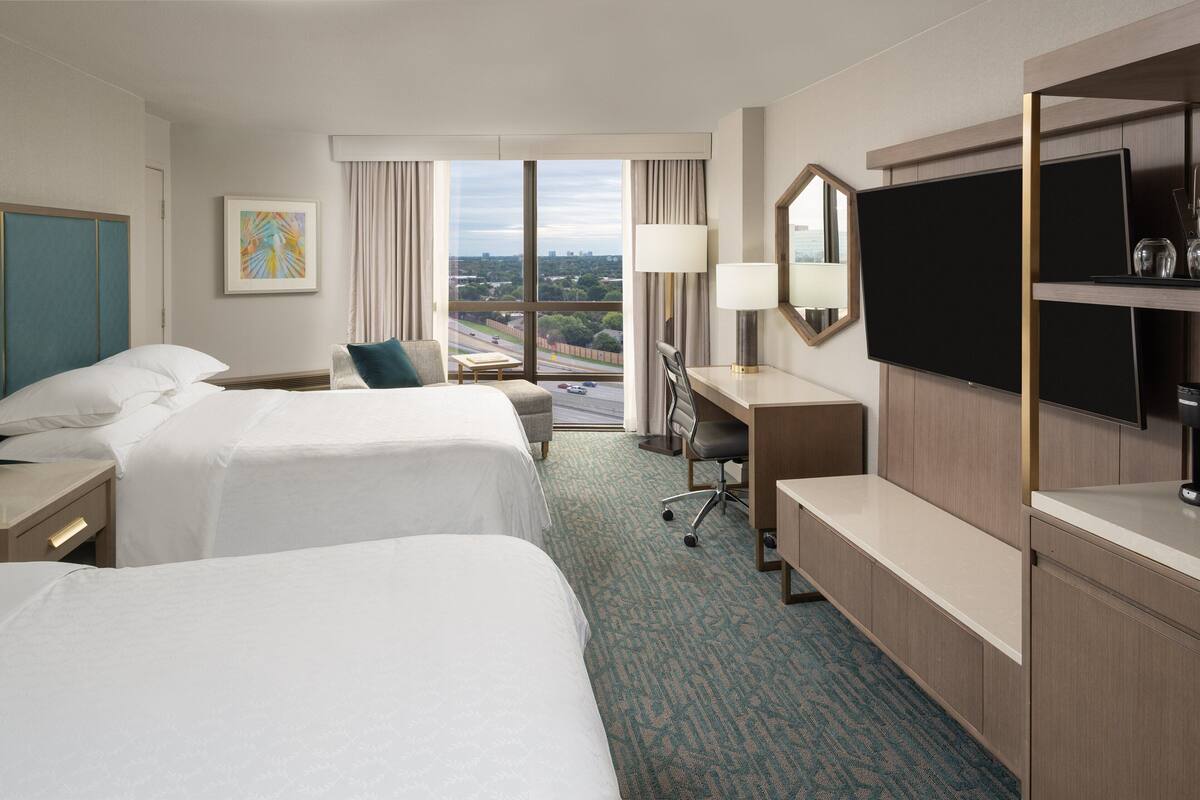 Habitación de hotel con dos camas, un escritorio, una silla, un televisor y un amplio ventanal con vistas a la ciudad.