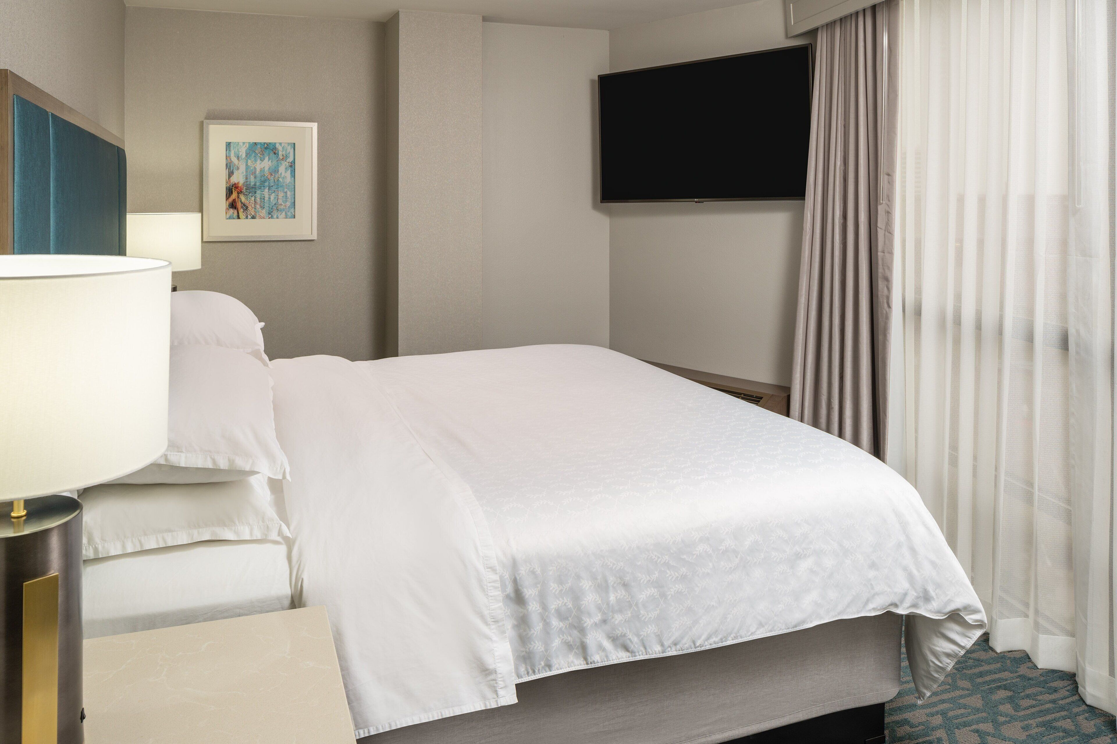 Habitación de hotel con cama, mesita de noche, lámpara, televisor de pantalla plana en la pared y ventana con cortinas.