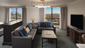 A modern hotel room with a sectional sofa, a flat-screen TV, a desk, and a large window offering a city view.