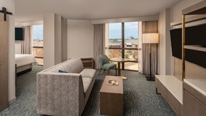 A hotel room with a bed, a sofa, a small table, a chair, a TV, and a large window with a city view.