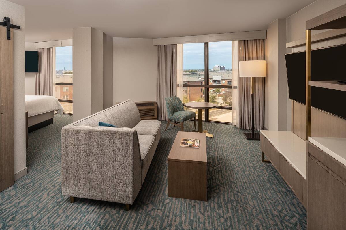 Habitación de hotel con cama, sofá, una mesita, una silla, televisor y un ventanal con vistas a la ciudad.