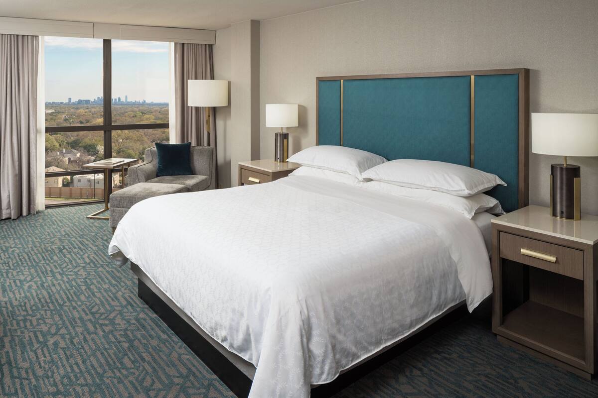 Habitación de hotel con una cama grande, dos mesitas de noche, una silla y vistas a la ciudad a través de amplios ventanales.