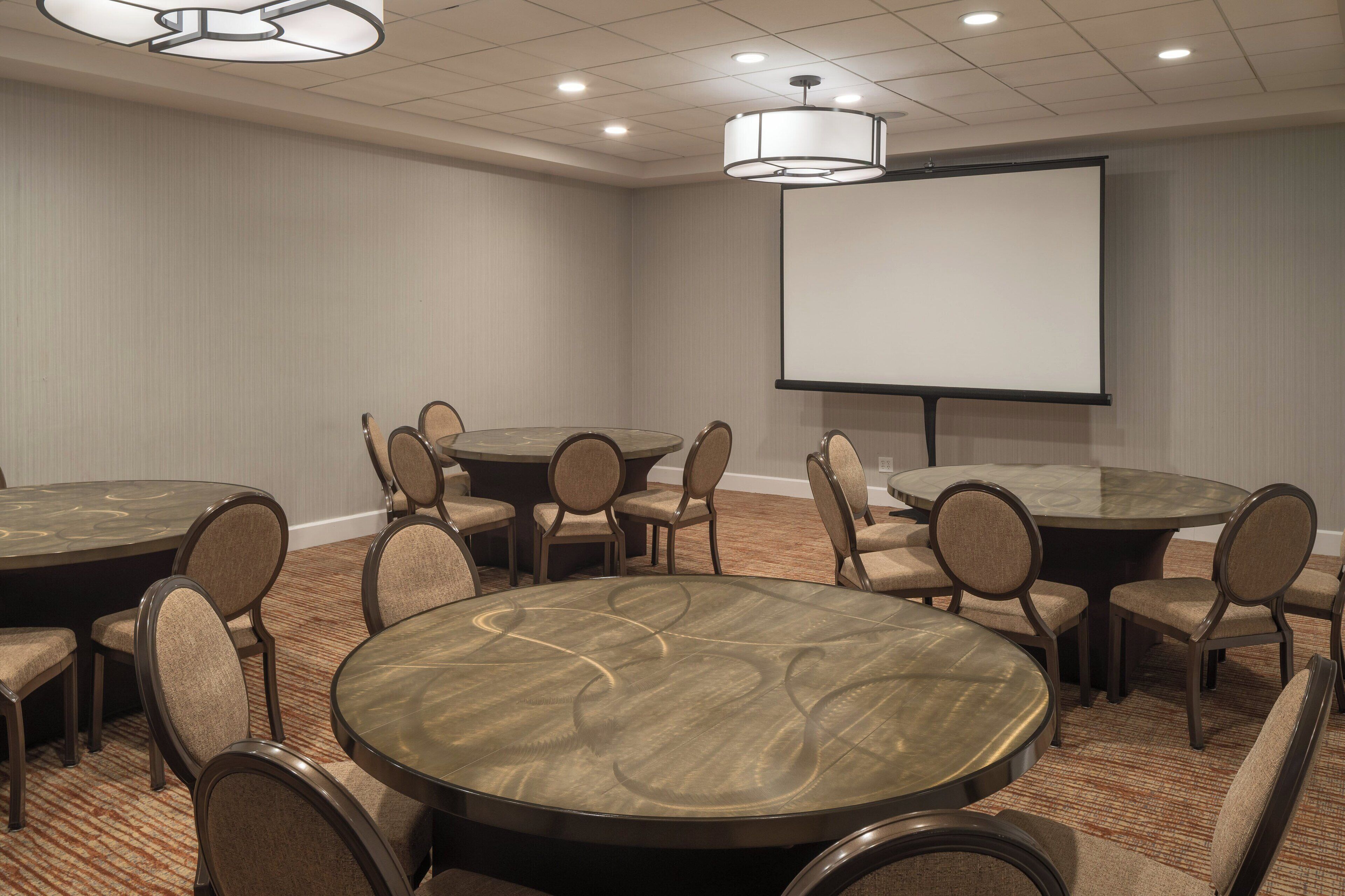 Un salón de conferencias con varias mesas redondas y sillas, una pantalla de proyector y piso alfombrado.