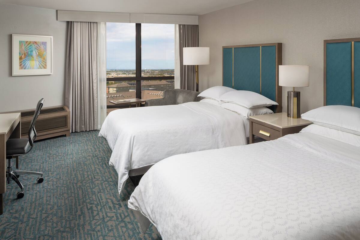 Habitación de hotel con dos camas, un escritorio, una silla y un ventanal con vistas a la ciudad.