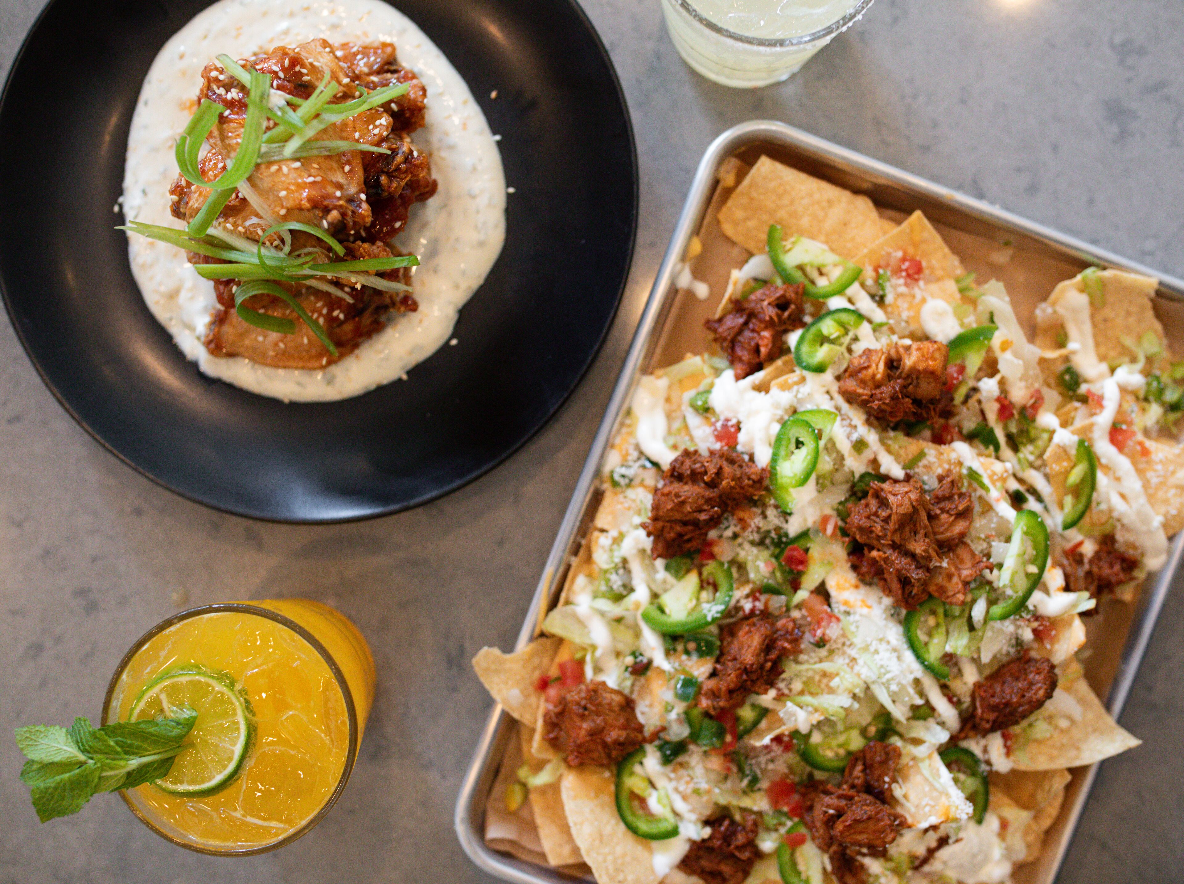 A tray of nachos topped with meat, cheese, and jalapeños, a bowl of nachos with a sauce and green onions, and a glass of orange juice with a lime slice.
