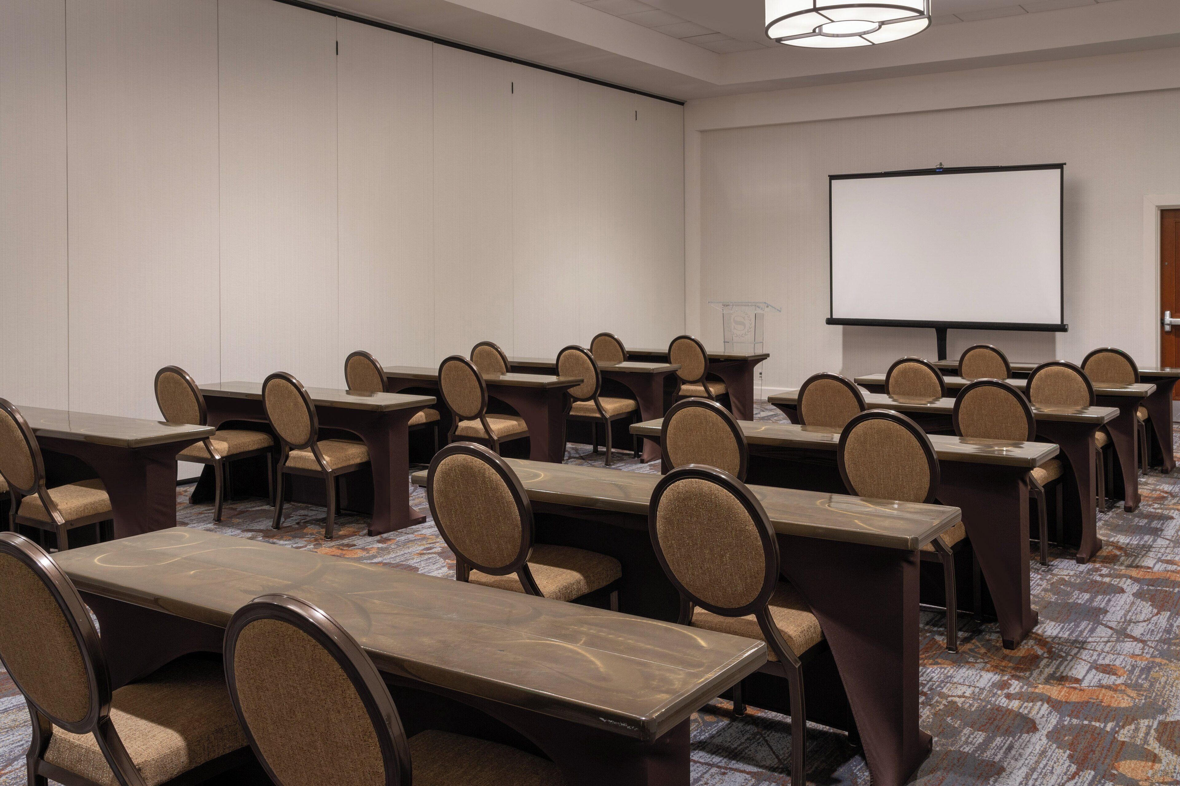 A conference room with multiple tables and chairs, a projector screen, and a carpeted floor.