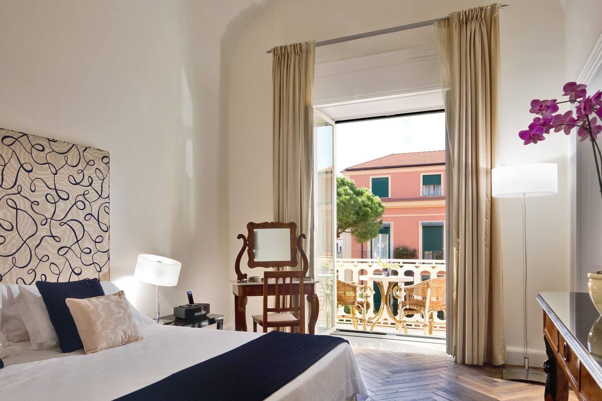 Habitación de hotel con cama, escritorio, silla, lámpara y vista al balcón exterior.