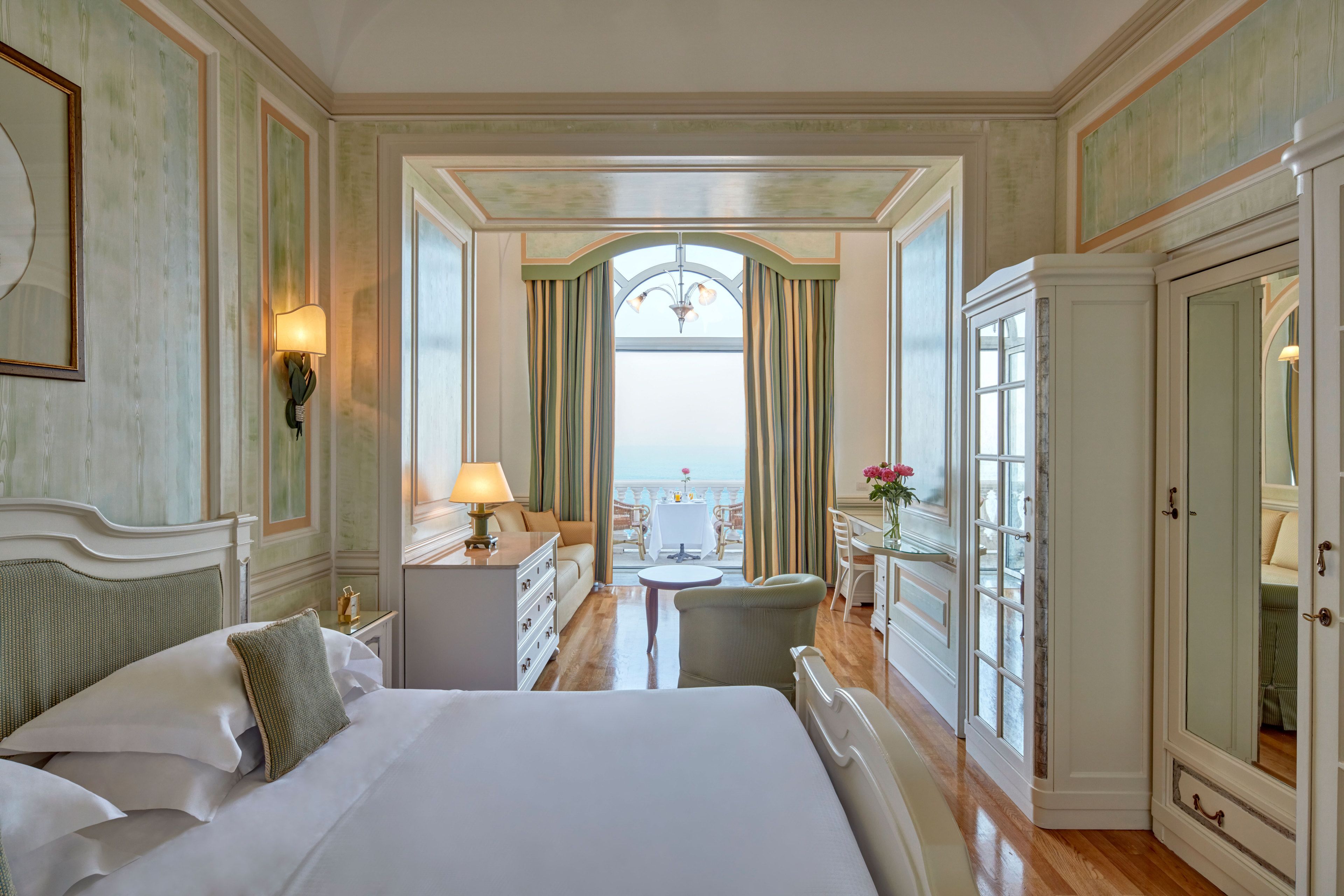 Une chambre d’hôtel avec un grand lit, un bureau, une chaise, une commode et une vue sur la mer depuis un balcon.