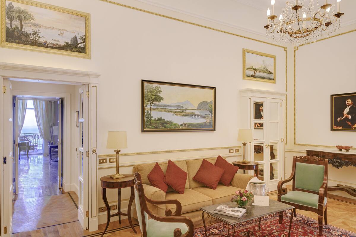 Un salón amplio con un sofá color beige, un sillón verde y una mesa de centro. Hay cuadros en las paredes y una araña en el techo.