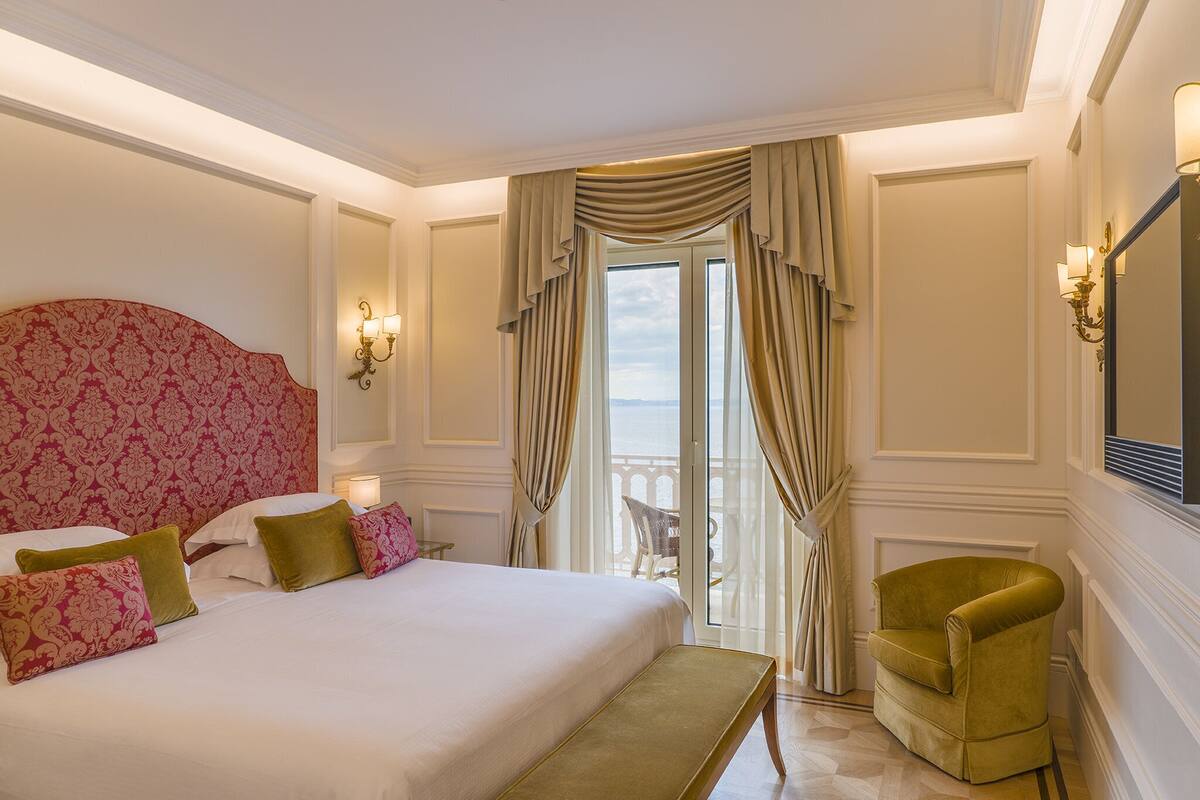 Habitación de hotel con una cama grande, un sillón verde y vistas al mar a través de la ventana.