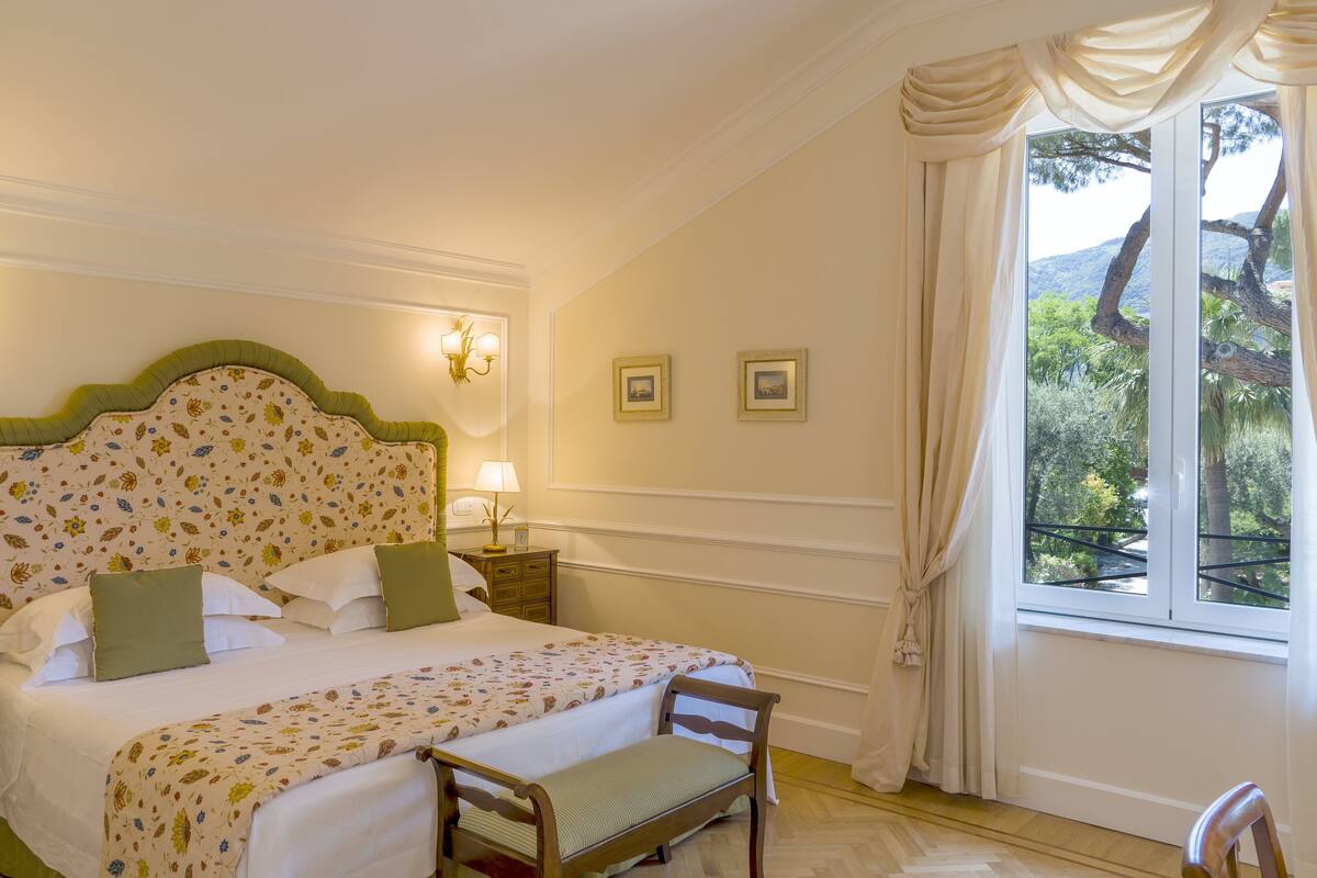 Un dormitorio con colcha estampada, cabecera de madera, un banco, dos mesitas de noche con lámparas y una ventana con cortinas.