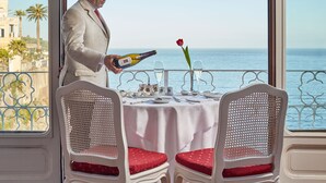 Un hombre con traje blanco sostiene una botella de champán, de pie junto a una mesa con dos sillas blancas y una rosa roja.