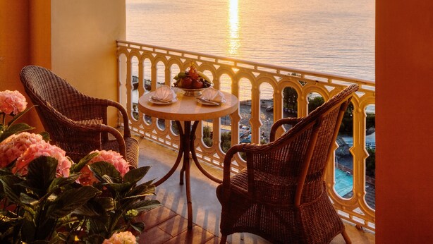 Un balcón con sillas de mimbre y una mesa, con vistas a un atardecer sobre el mar.