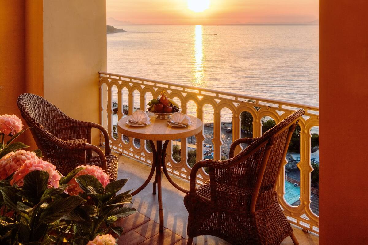 Un balcón con sillas de mimbre y una mesa, con vistas a un atardecer sobre el mar.