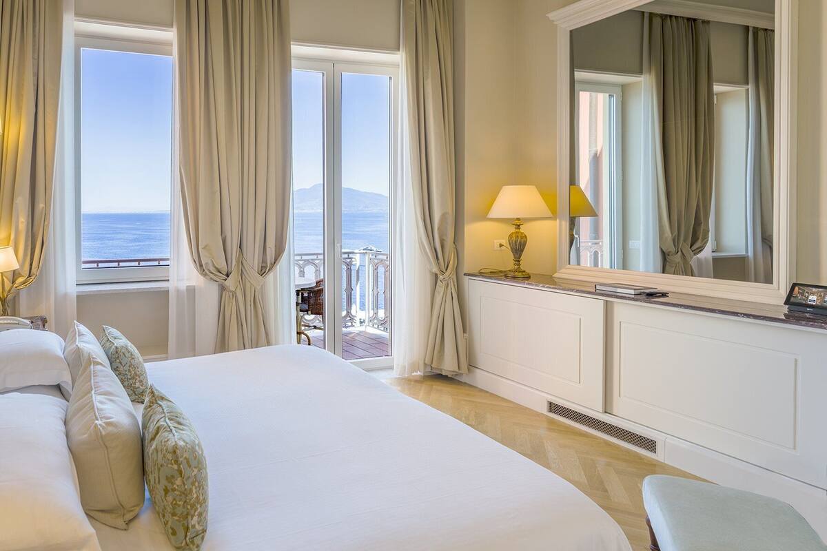 Habitación de hotel con una cama grande, un escritorio con lámpara, un espejo y vistas al mar a través de amplias ventanas.