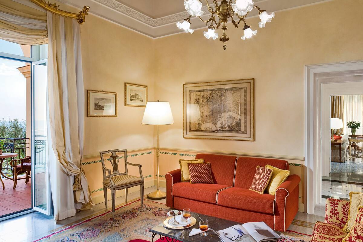Un salón espacioso con un sofá rojo, una mesa de comedor con sillas, una araña y un ventanal con cortinas.