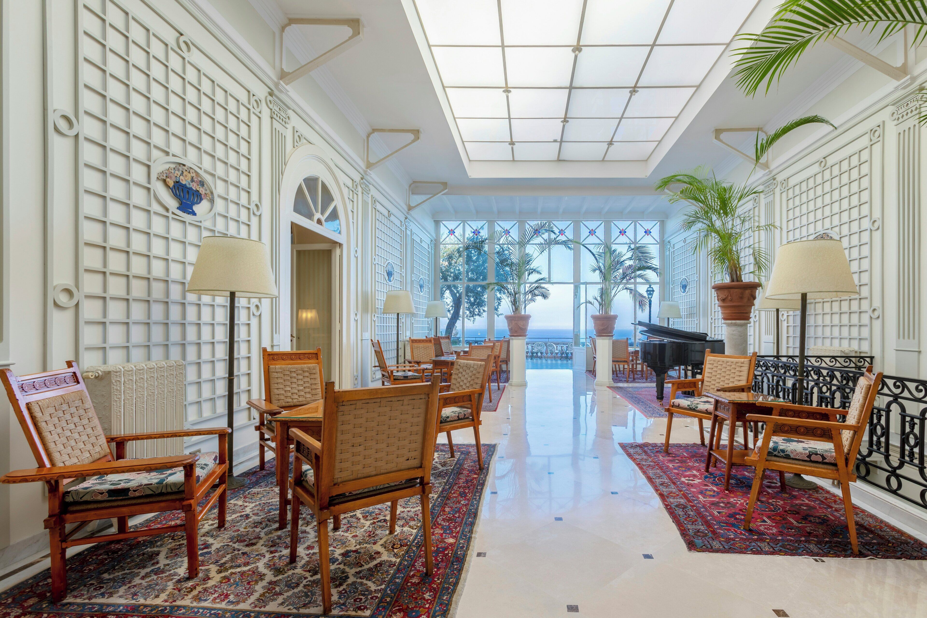 A spacious lobby with a grand piano, wooden chairs, a patterned rug, and a view of palm trees.