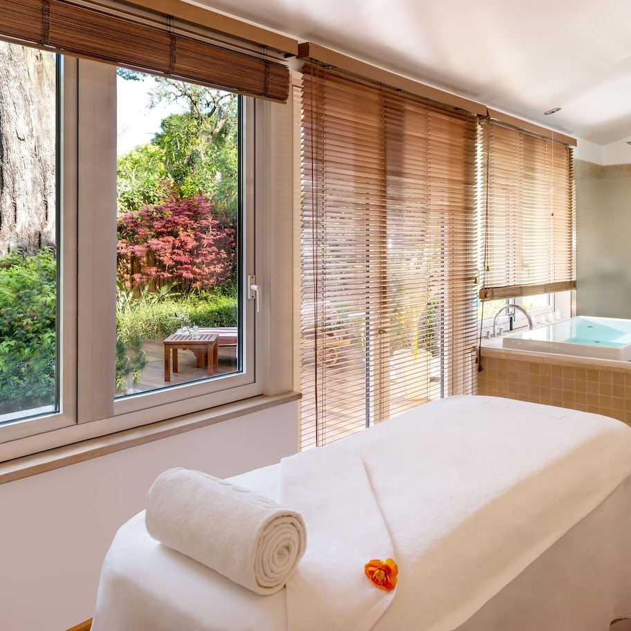 A spa room with a massage table, a bathtub, and a view of a garden through large windows.