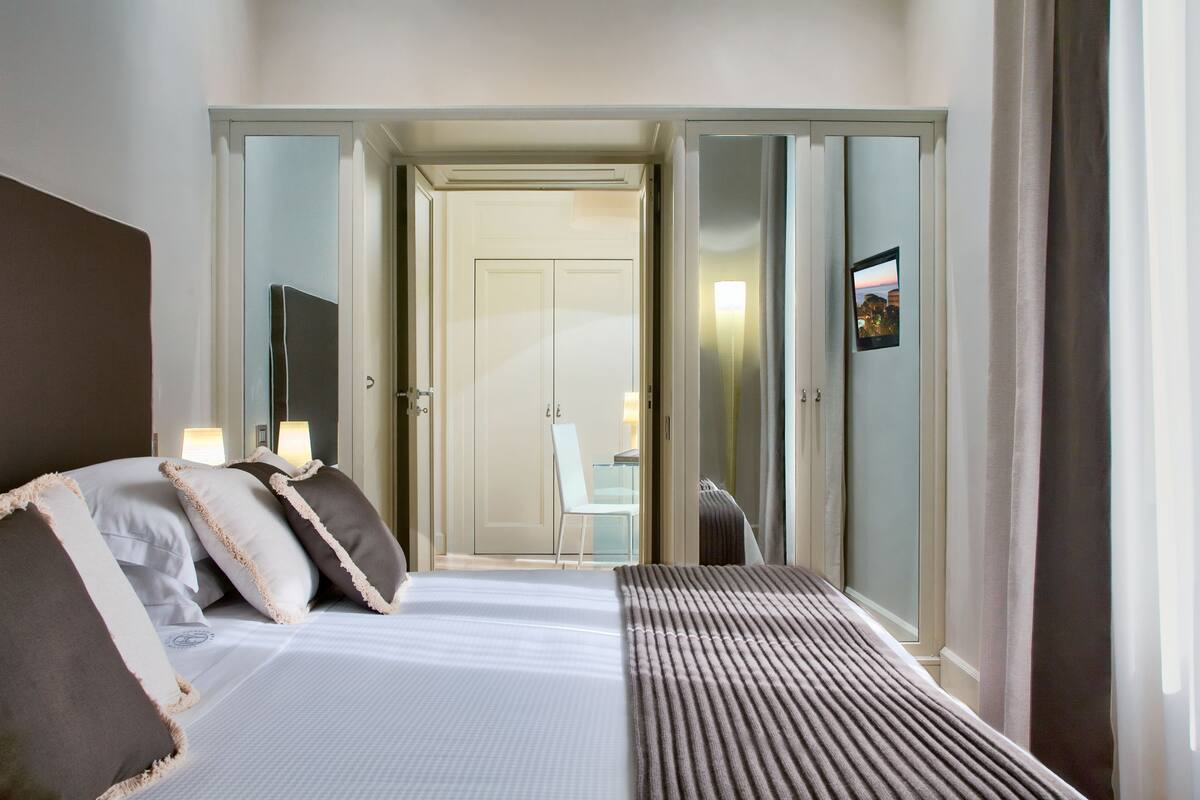 Habitación de hotel con cama, lámparas de noche, un espejo y vista al pasillo.