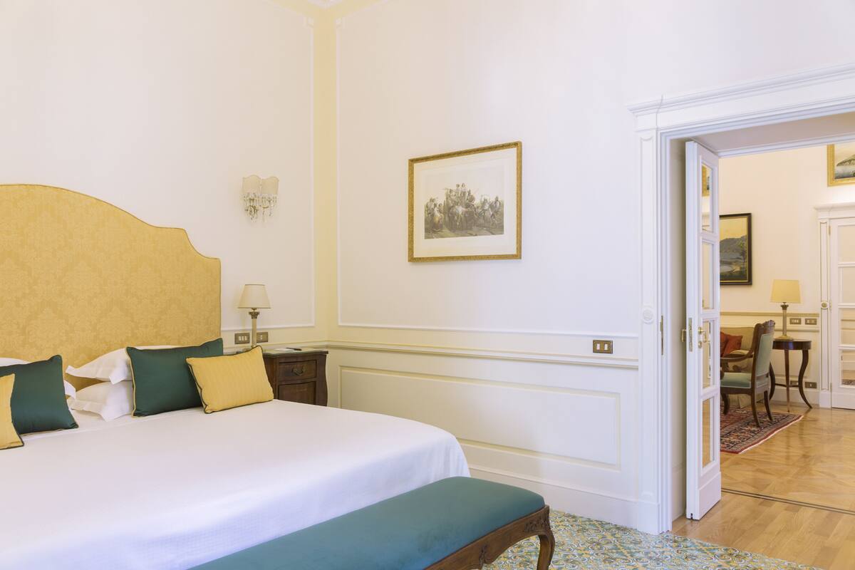 Habitación de hotel con una cama grande, una mesita de noche, una silla y una puerta que da a otra habitación.